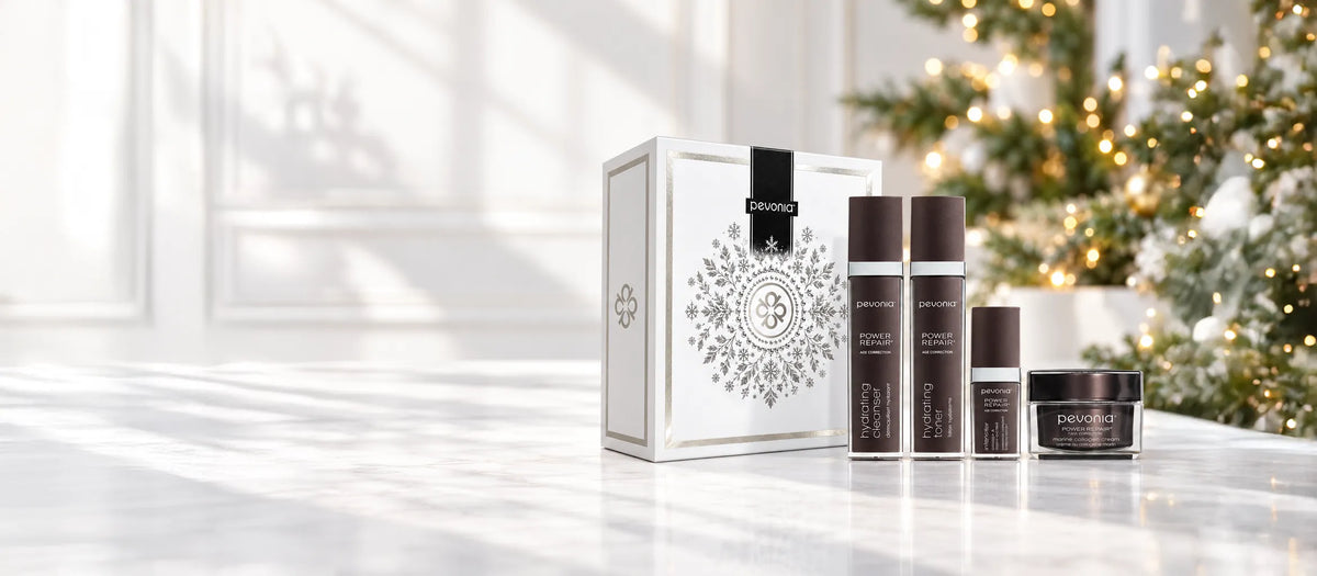 Beauty Collective Christmas Gift Collection with a white decorative box arranged on a light marble surface. A softly blurred Christmas tree with warm lights appears in the background