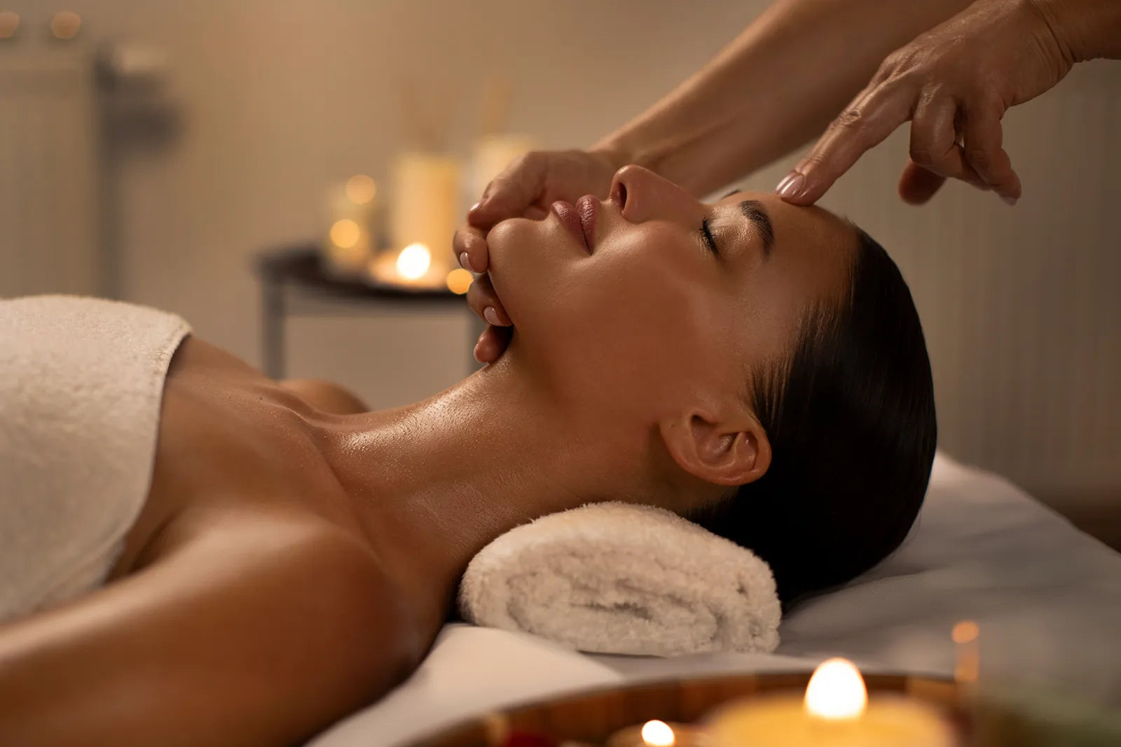 A person with dark hair lies on a massage table with a towel under their head, eyes closed, while receiving a relaxing facial massage in a softly lit spa setting with candles.