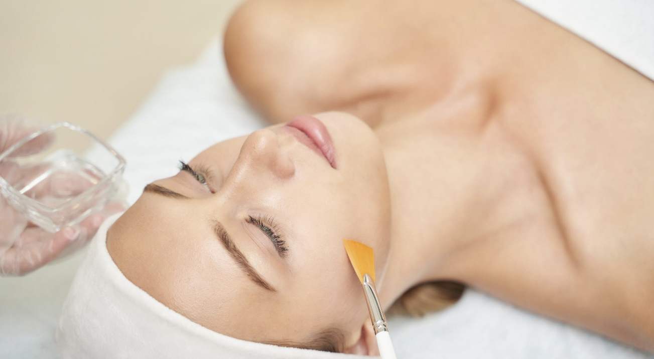 A woman with a headband lies on a spa bed with her eyes closed while a brush applies a facial treatment to her cheek. A hand holds a small glass container nearby. The atmosphere appears calm and relaxing.