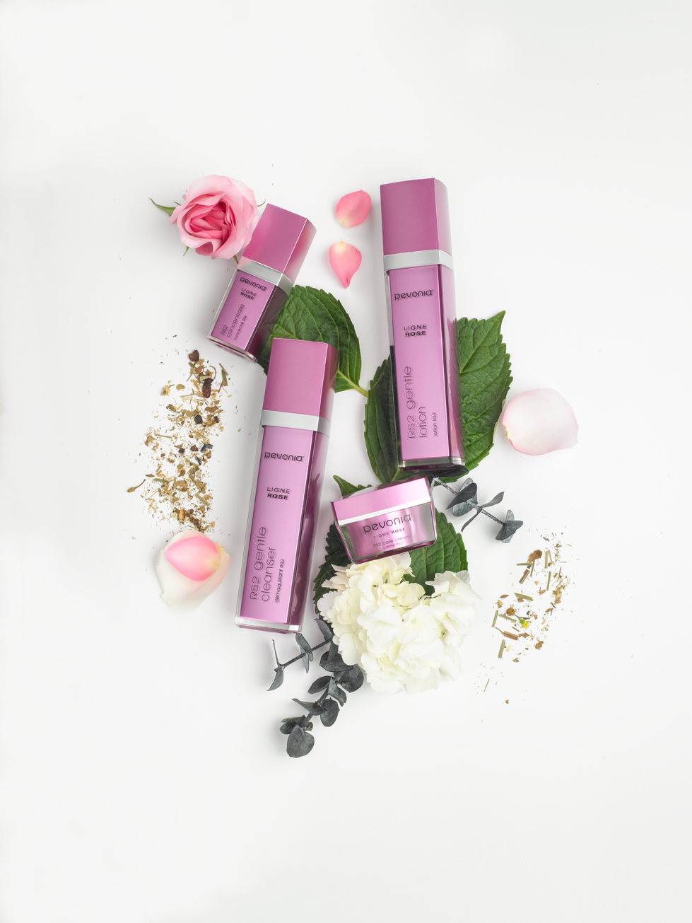 Four pink skincare bottles are arranged on a white background, surrounded by pink and white flowers, green leaves, petals, and gold flecks, creating an elegant and fresh display.